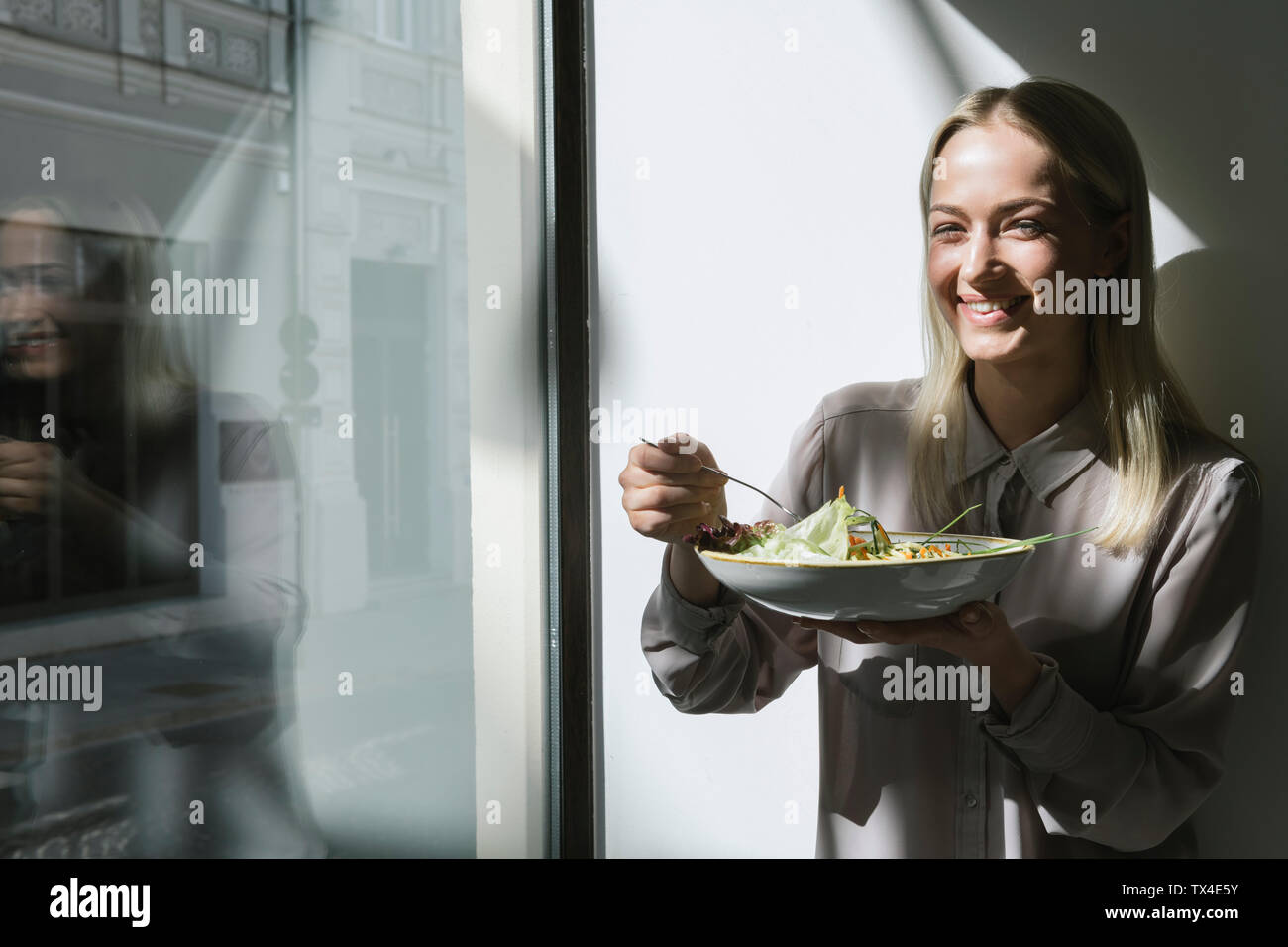 Lächelnde junge Frau steht am Fenster in der Sonne essen einen Salat Stockfoto