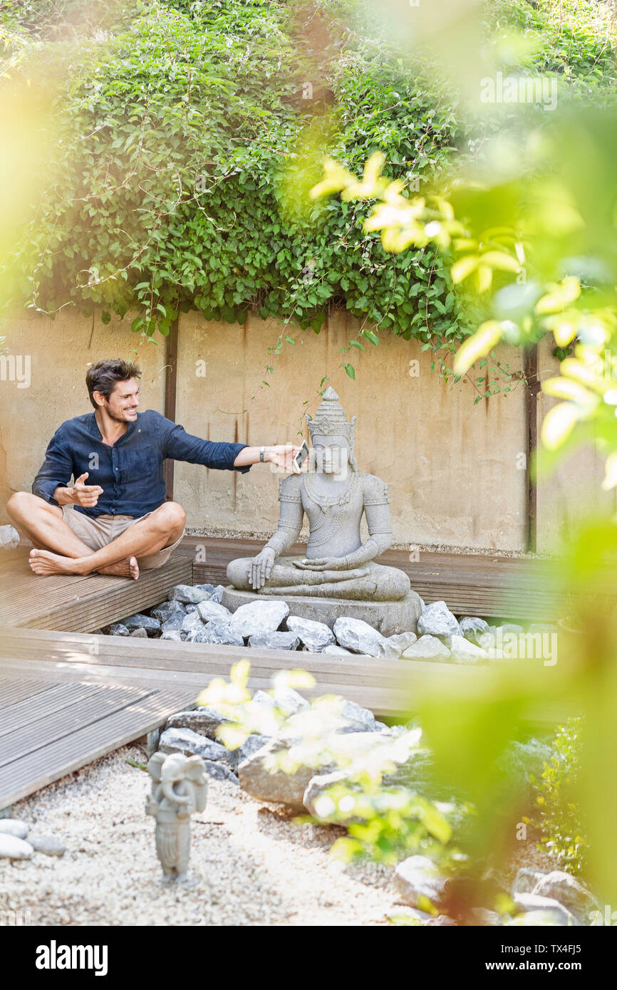 Mann mit überkreuzten Beinen neben Buddha Statue in einem Zen-garten mit Smartphone Stockfoto