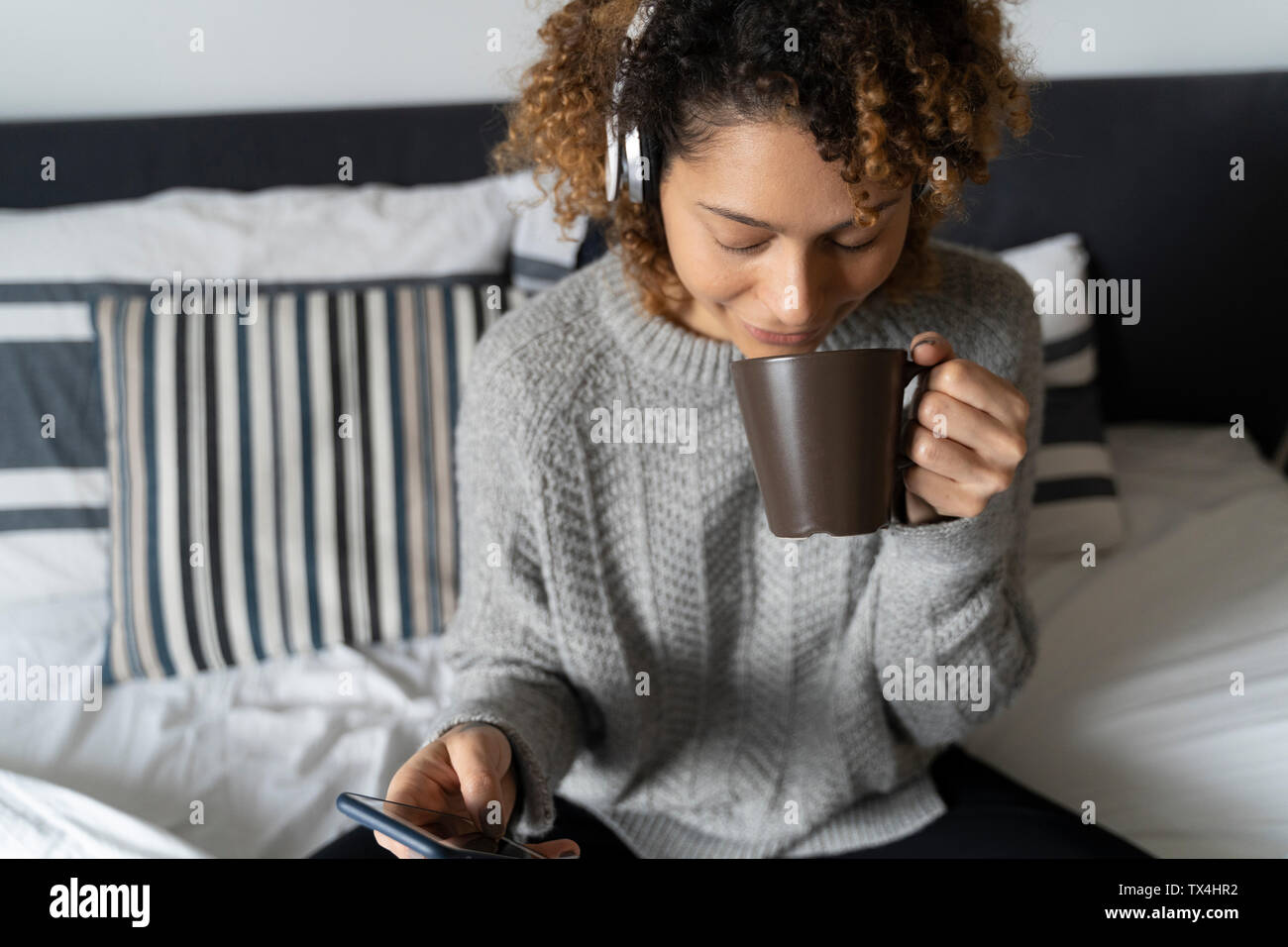 Frau sitzt auf dem Bett, Kaffee trinken, Musik hören mit Kopfhörern und Smartphone Stockfoto