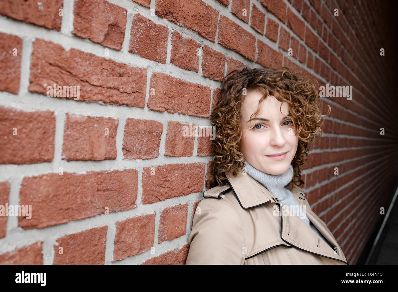 Portrait von Frau mit lockigem Haar beigefarbenen Trenchcoat gegen die Mauer gelehnt Stockfoto