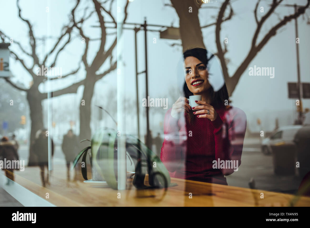 Lächelnde junge Frau mit Tasse Kaffee hinter Fensterglas in einem Cafe Stockfoto