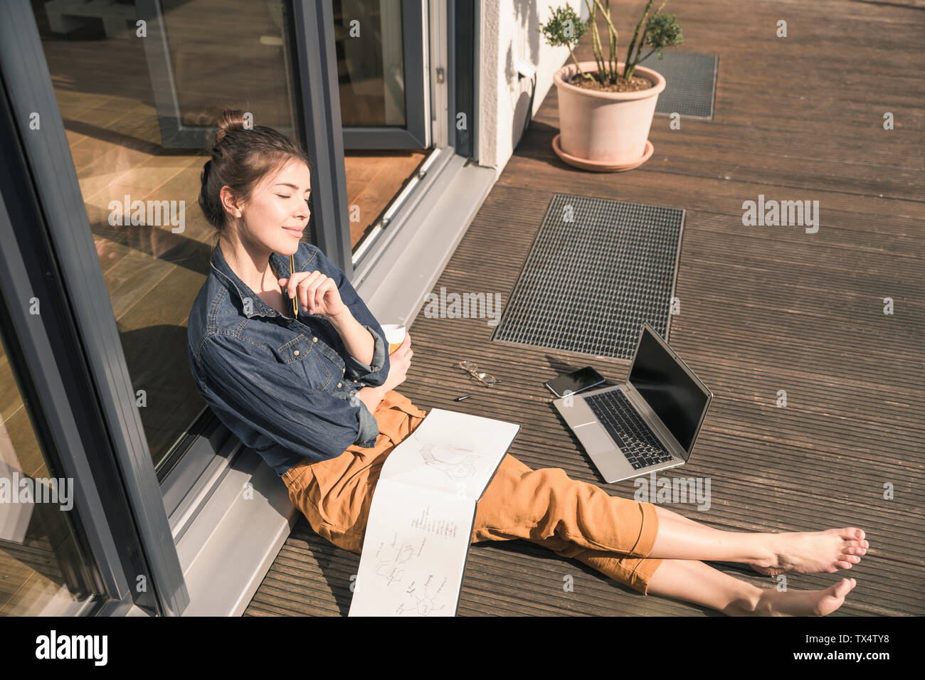Junge Frau mit geschlossenen Augen sitzen auf der Terrasse zu Hause mit Laptop und Buch Stockfoto