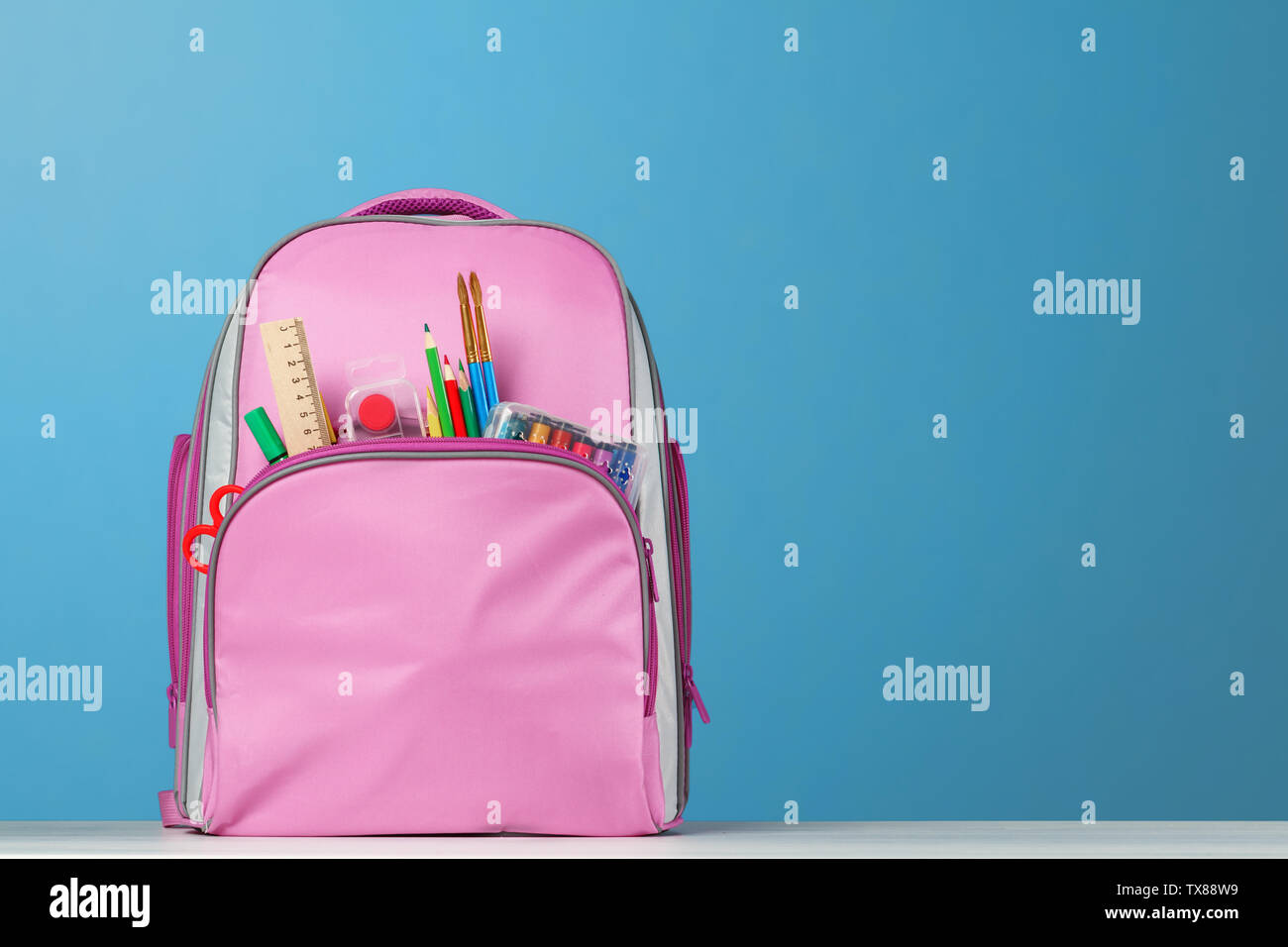 Rosa Rucksack mit Bürobedarf auf dem Tisch auf einem blauen Hintergrund. Zurück zu Schule. Stockfoto