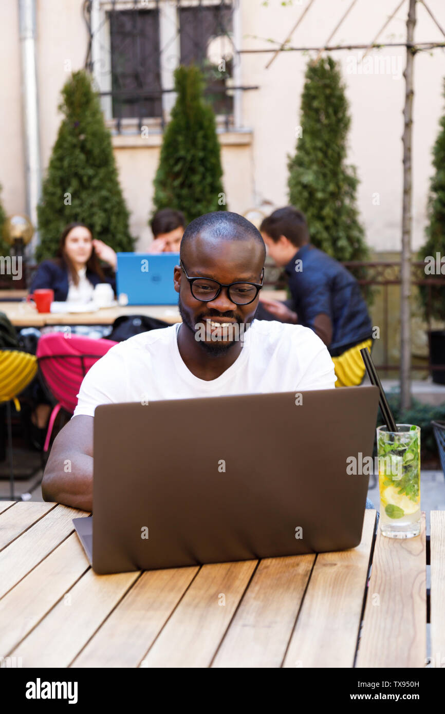 Afrikanische hübscher Junge sitzt mit mohito am Café im Freien und die Arbeit mit Laptop Stockfoto