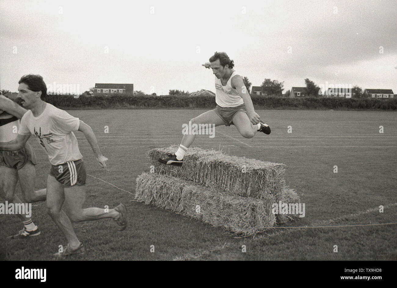 1980 s, amateur männlicher Athletik Teil in einem Hindernislauf rennen Außerhalb, in einem Feld, die mit einer der Wettbewerber über ein Hindernis von Strohballen, England, UK springen. Stockfoto