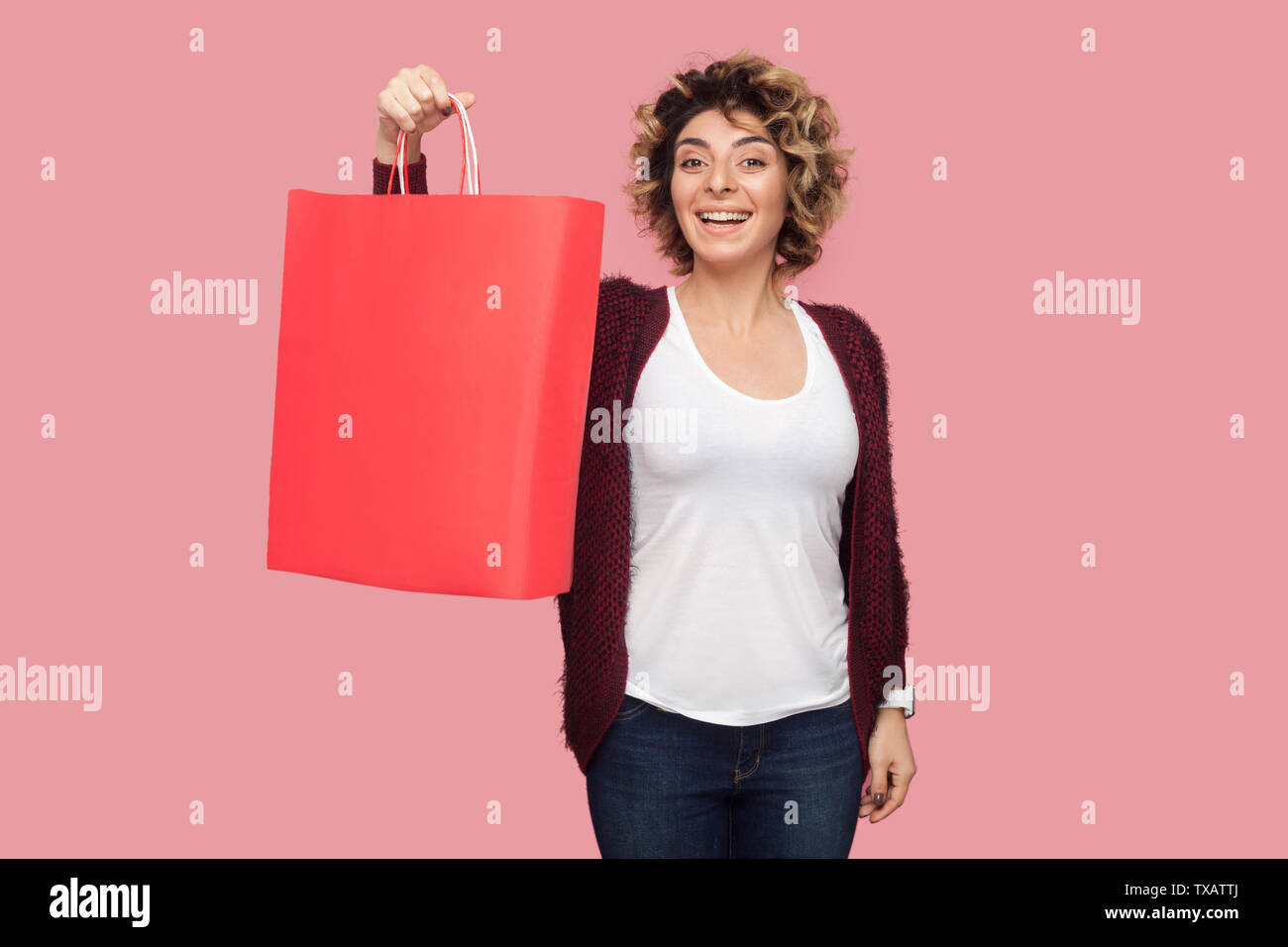 Wow! Moderne junge Frau im blauen Hemd mit curlty Frisur stehen und mit Einkaufstüten und toothy Lächeln zufrieden, bei Kamera schaut. Studio s Stockfoto