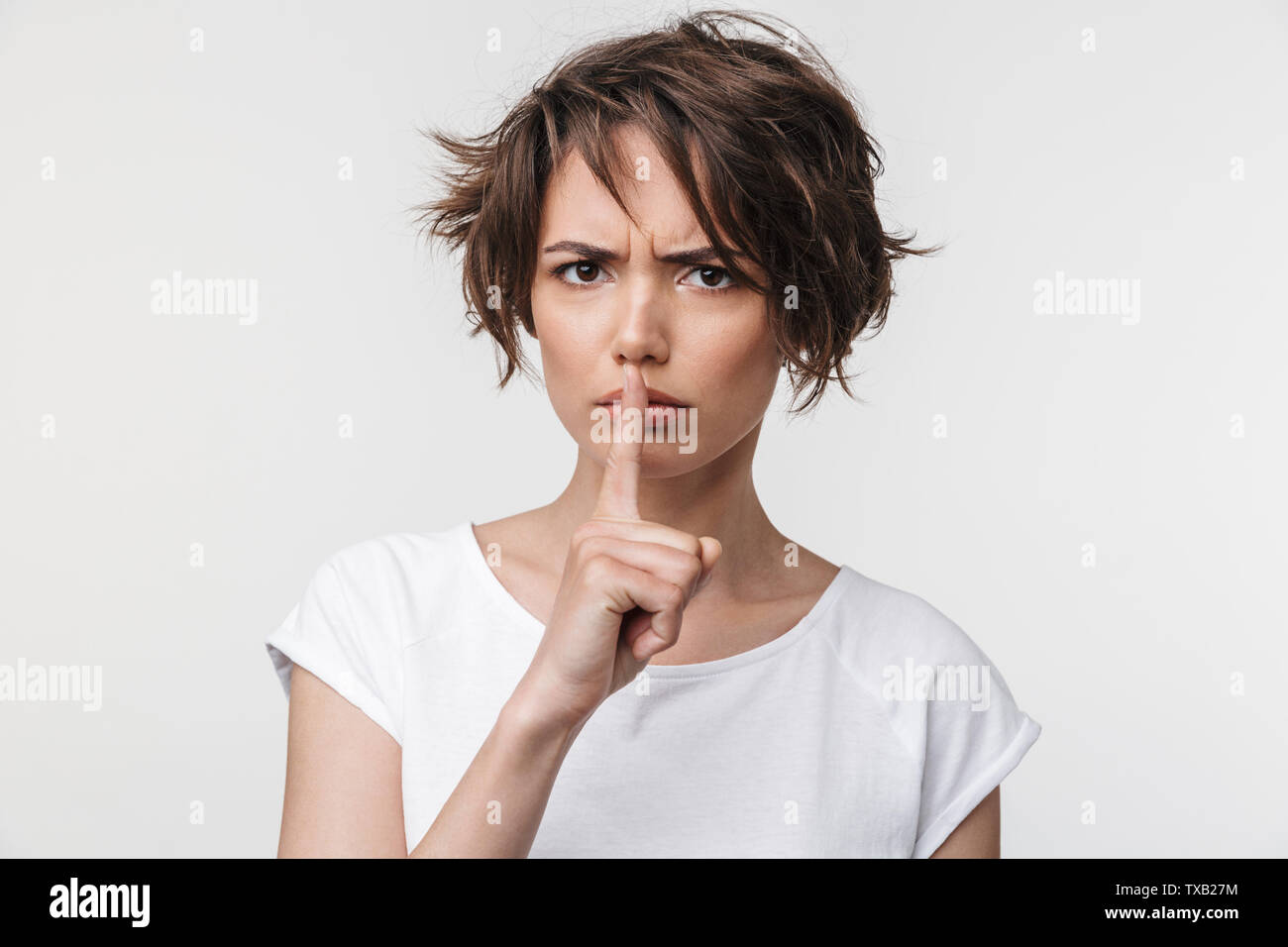 Portrait von wütende Frau mit kurzen braunen Haaren in Basic T-Shirt mit Zeigefinger auf die Lippen, während auf weißem Hintergrund stehend Stockfoto