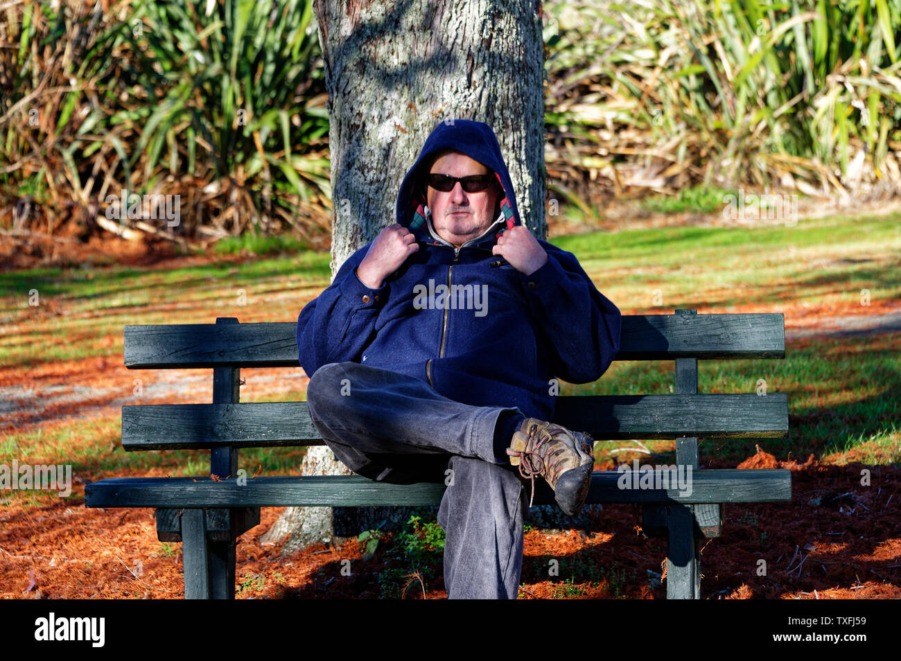 Ein Mann sitzt in einem Park mit seiner Sonnenbrille auf und seine Haube Stockfoto
