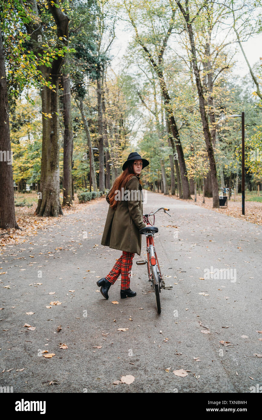Junge Frau mit langen roten Haaren schaut über ihre Schulter, während Fahrrad schieben im Herbst Park, volle Länge, Florenz, Toskana, Italien Stockfoto