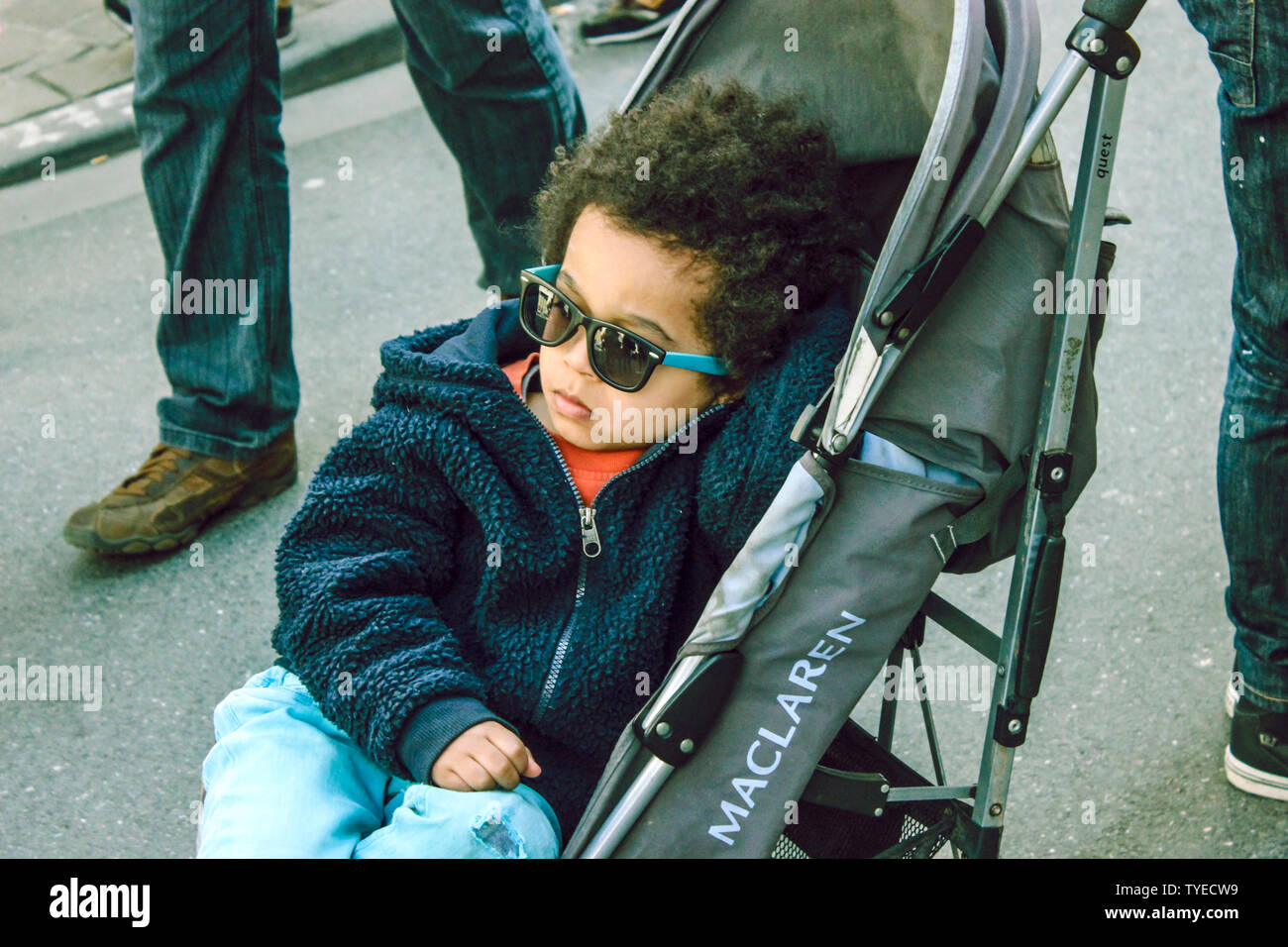 Juni 2019, Brüssel, Belgien. In seinem Kinderwagen/Schiebestuhl trägt er eine schöne und stilvolle Sonnenbrille. Coole süße schwarze Kid w blaue Sonnenbrille und lockiges Haar Stockfoto