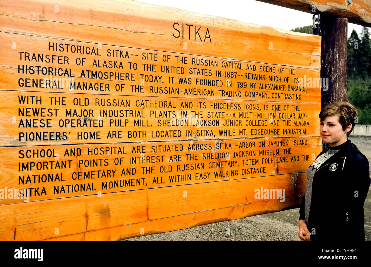 Ein Besucher in Alaska, USA, liest eine hölzerne Zeichen, die eine kurze Geschichte von Sitka, die neue Erzengel genannt wurde, wenn es war die Hauptstadt der Russischen Hoheitsgebiet vor dem Russland Alaska in die Vereinigten Staaten im Jahr 1867 verkauft. Zu den Sehenswürdigkeiten der Russischen Sitkas Erbes ist die 1848 Saint Michael's russisch-orthodoxe Kathedrale, die durch einen Brand im Jahre 1966 zerstört, aber wieder aufgebaut als Replik mit seinem zwiebelförmigen Kuppel und nadelförmige Glockenturm gekrönt von goldene Kreuze. Viele der Russischen ära Icons wurden von der Feuersbrunst gespeichert und sind auf dem Display im Inneren der Kirche. Stockfoto