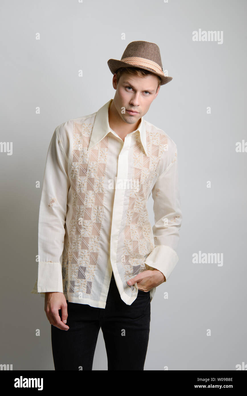 Ein gutaussehender kaukasischer Mann mit braunem Haar posiert in einem cremefarbenen Vintage 70s-Hemd und trägt einen braunen Fedora-Hut. Stockfoto