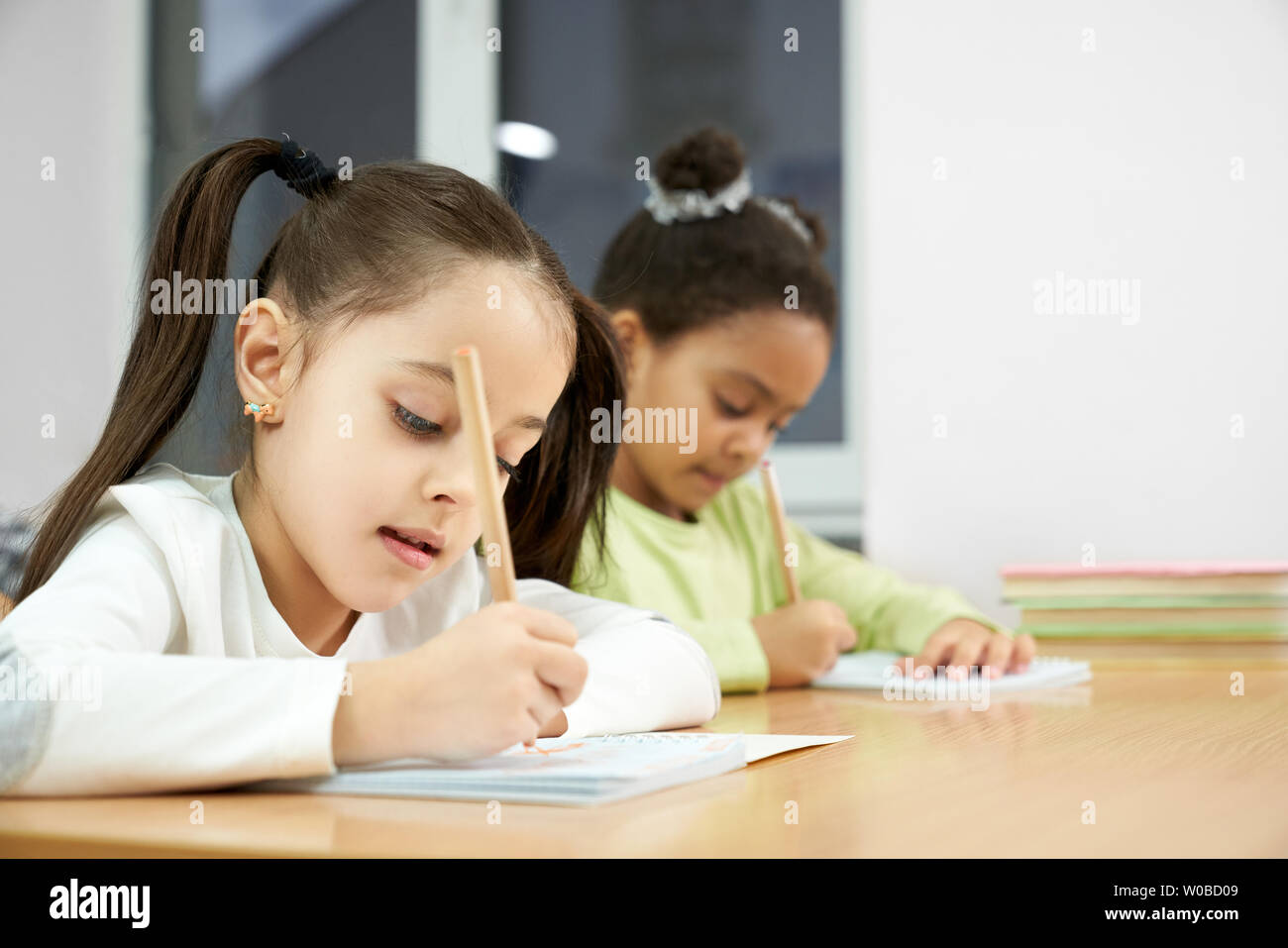 Freundliche hübsche Mädchen der Grundschule schreiben in Copybook mit Bleistift. Niedlich, konzentrierten Schülerinnen im Klassenzimmer sitzen, in dem Notebook, bei Lektion posieren. Stockfoto