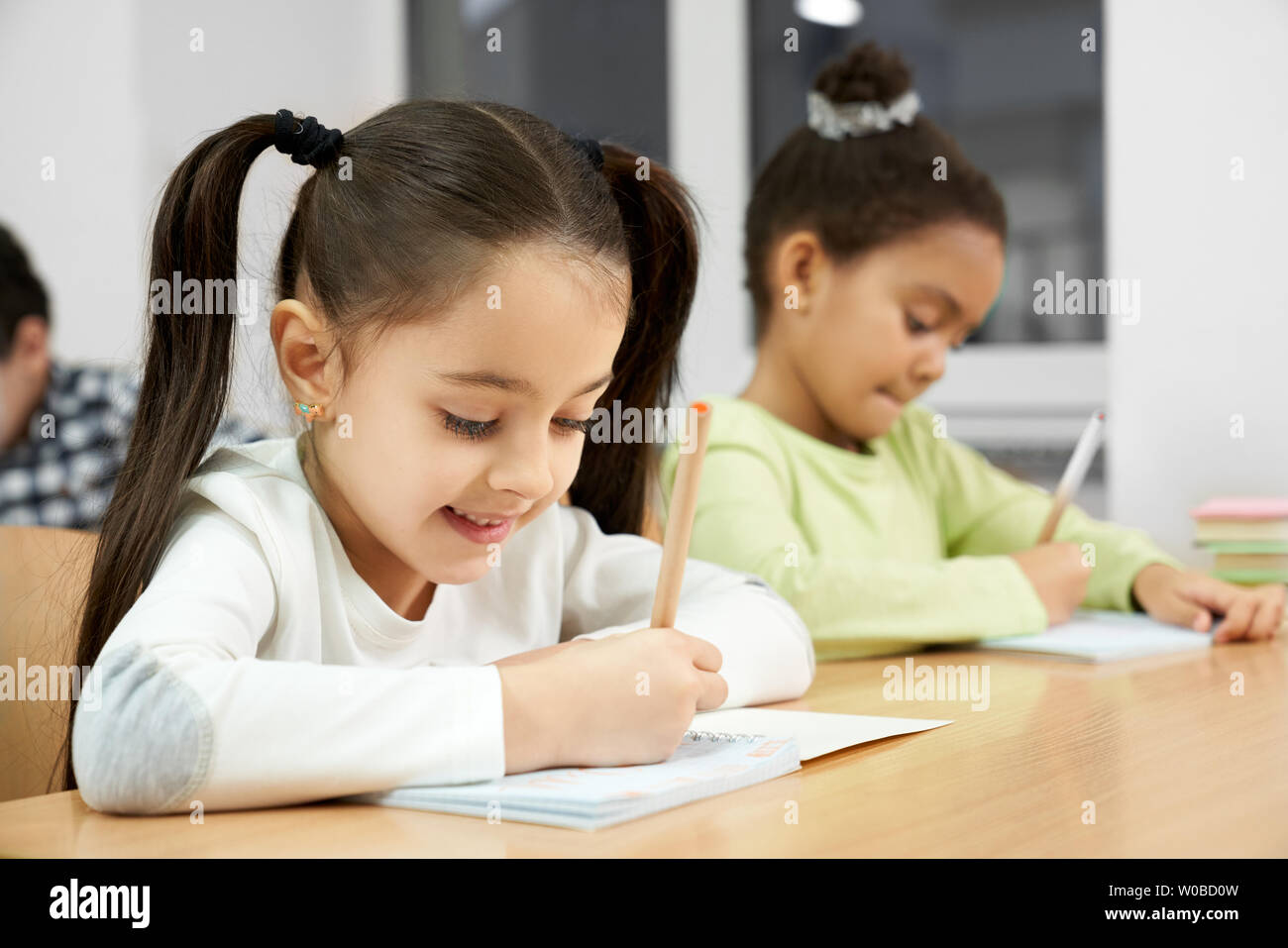 Schön, glücklich, Schülerinnen der Grundschule am Schreibtisch im Klassenzimmer sitzen, Bleistifte und sorgfältig Schreiben in Copybook. Mädchen auf der Suche im Notebook, posierend, lächelnd. Stockfoto