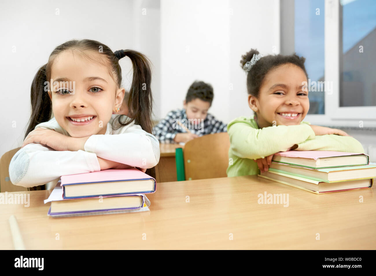 Lustig, hübsch, Schülerinnen am Schreibtisch im Klassenzimmer sitzen, lächelte und lehnte sich auf Bücher. Fröhliche Schüler, die Mitschüler der Grundschule auf Kamera posieren. Stockfoto