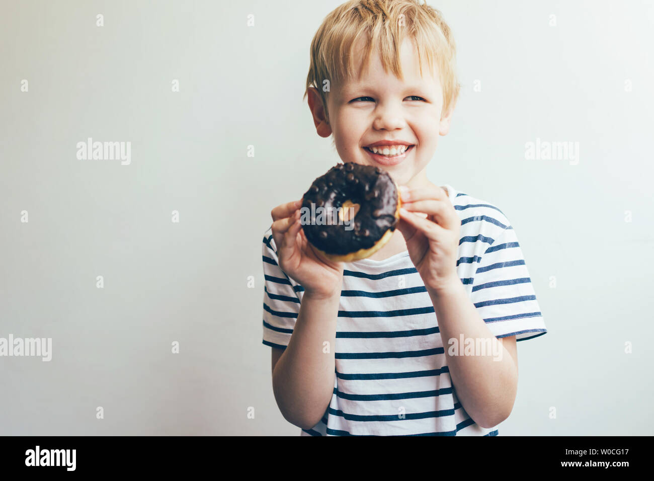 Kaukasische blonde Kind 5-6 Jahre alt in einem gestreiften Casual T-Shirt auf einem weißen Hintergrund mit einem leckeren Schokolade Donut in der Hand und lächelt h Stockfoto