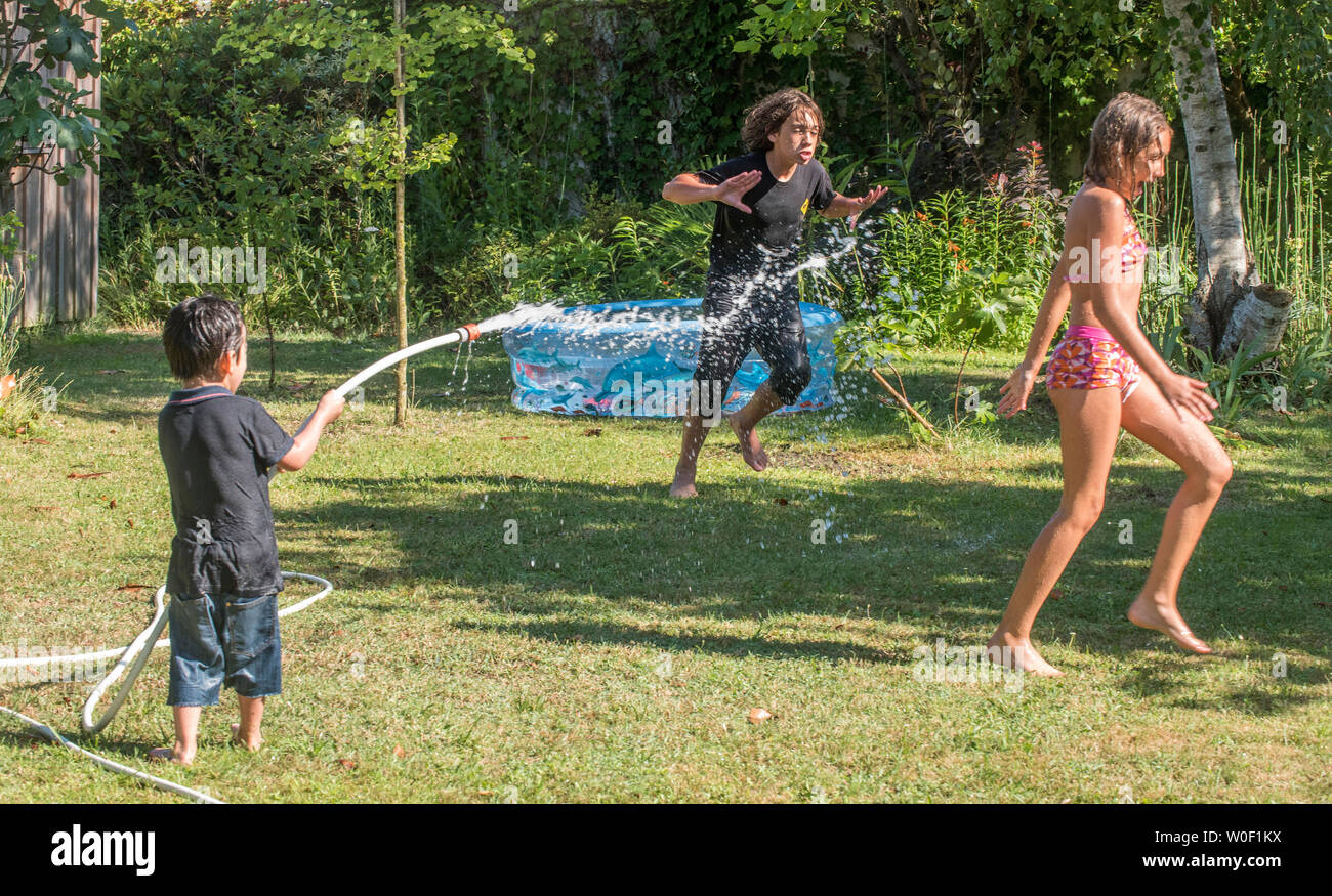 Zwei Jungen, der 4 Jahre alt ist und 12 Jahre alt und ein 10-jähriges Mädchen spielen mit einem Schlauch im Garten Stockfoto