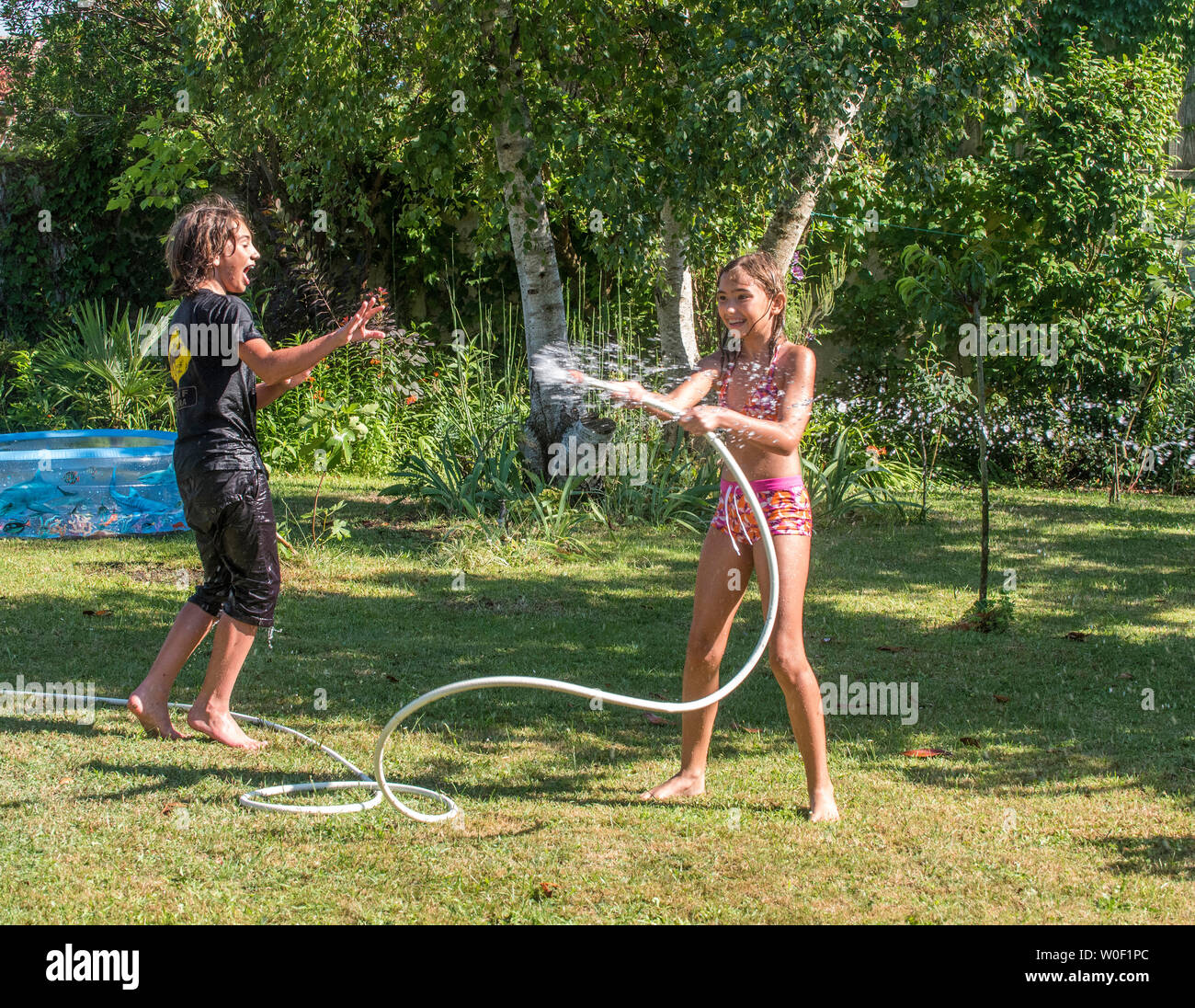 Zwei Jungen, der 4 Jahre alt ist und 12 Jahre alt und ein 10-jähriges Mädchen spielen mit einem Schlauch im Garten Stockfoto