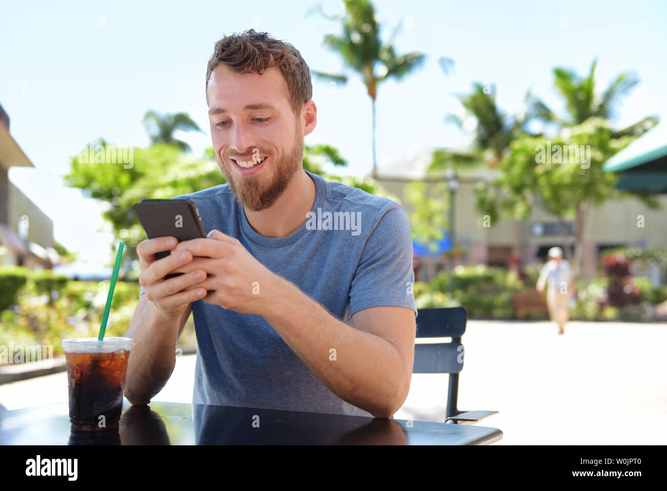 Mann auf Cafe mit Smart Phone app Text Messaging sms trinken Eiskaffee im Sommer. Hübscher junger legerer Mann mit Smartphone lächelt glücklich Sitzen im Freien. Urban Mann in seinen 20ern. Stockfoto