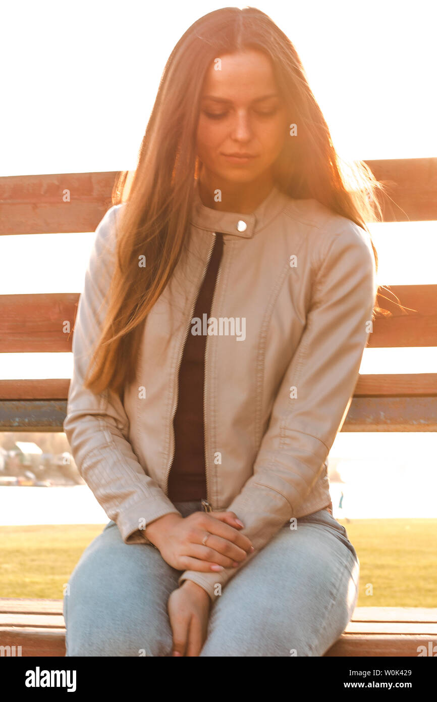 Schöne Mädchen mit langem Haar sitzt gegenüber dem Sonnenlicht in beige Lederjacke und Jeans. Nach unten schauen. Stockfoto