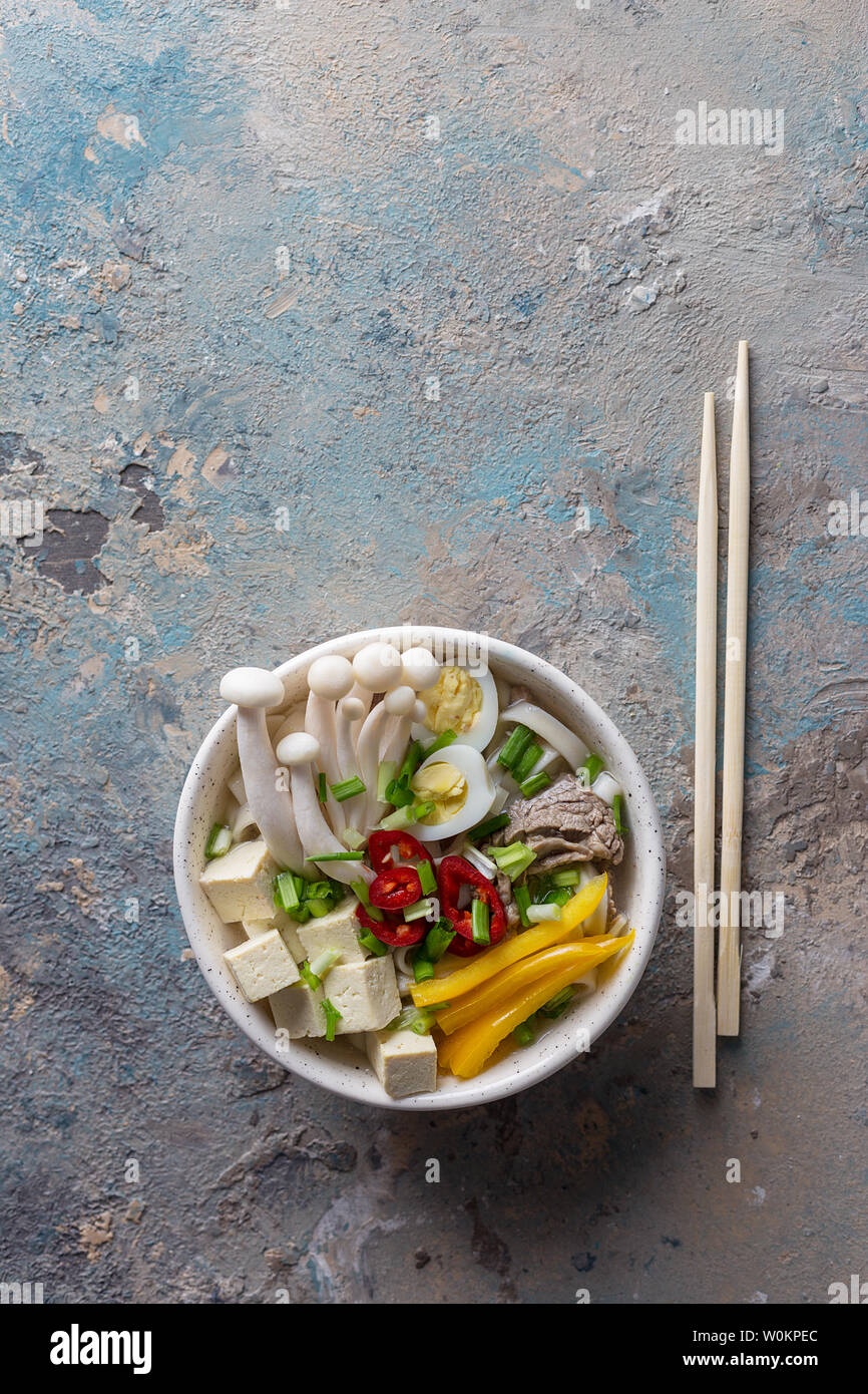 Heiße Suppe mit Nudeln, Chili, Tofu, grüne Zwiebeln und Pilze in Schale mit Stöcken auf konkrete blauen Hintergrund. Vertikal oben vie Stockfoto