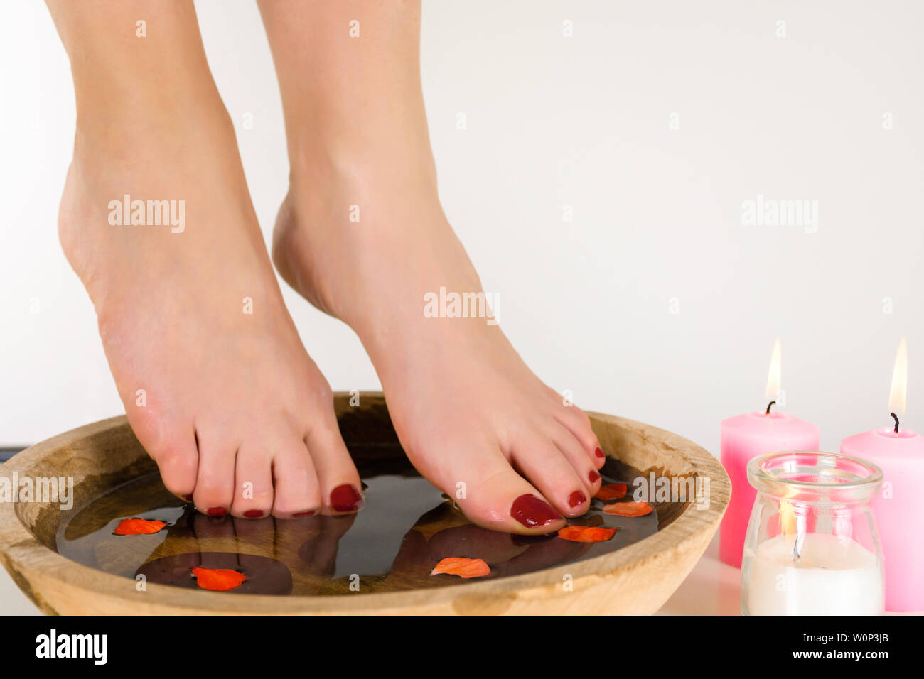 Weibliche Füße in eine Schüssel mit Wasser und Holz rosa Kerzen brennen auf dem Fußboden, weißen Hintergrund. Burgund Farbe auf den Nägeln Pediküre. Nahaufnahme Stockfoto