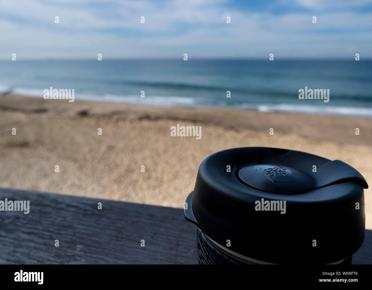 NEWCASTLE, AUSTRALIEN - 16. Juni 2019 in der Nähe von einem Glas Keepcup mit schwarzen Trimmen, gefüllt mit Kaffee, auf einer Bank mit den Pazifischen Ozean in t Stockfoto