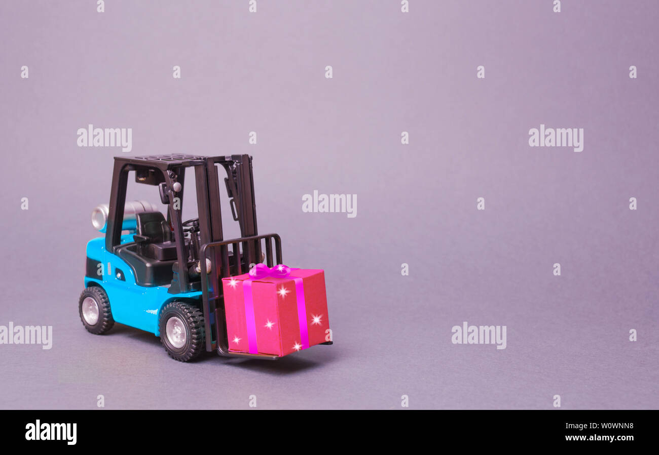 Blau Gabelstapler trägt eine rosa Geschenkbox mit Schleife. Kauf und Lieferung eines präsentieren. Einzelhandel, Rabatte und Wettbewerbe. Wettbewerb Werbeaktionen. Erhöht Stockfoto