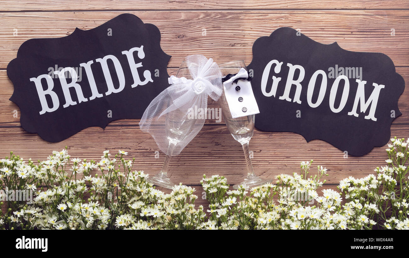 Champagner Glas in Wedding Dress mit Braut und Bräutigam text Zeichen auf hölzernen Hintergrund dekoriert mit kleinen weißen Blüten, Vintage Style. Hochzeit Schild con Stockfoto