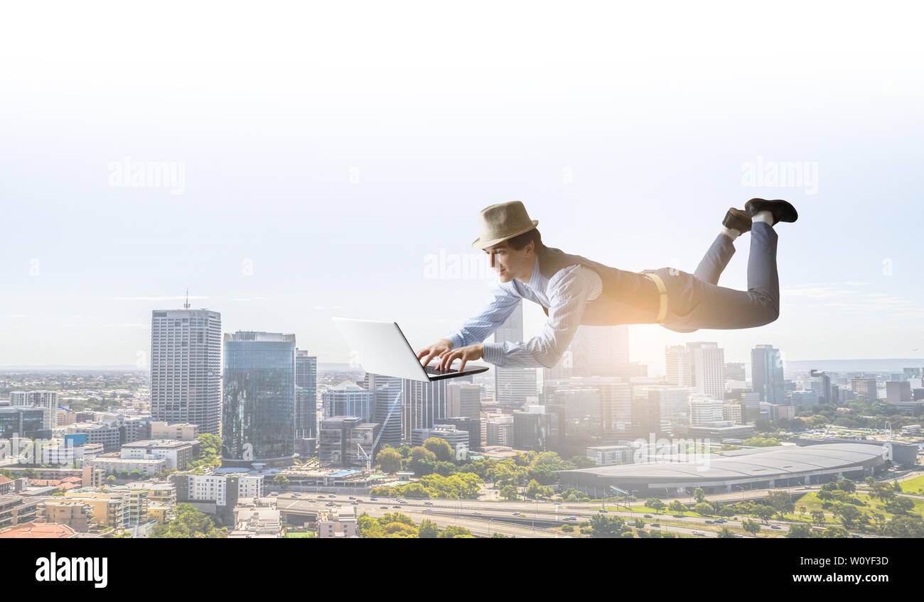 Junger Mann in Hut und legere Kleidung schwebend über Stadt mit Laptop. Mixed Media Stockfoto