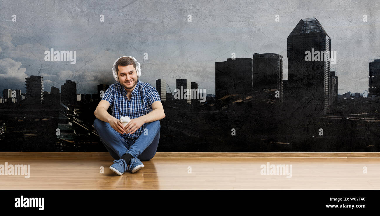 Junge fröhliche Mann in Freizeitkleidung sitzt auf dem Boden mit Kopfhörern auf dem Kopf. Mixed Media Stockfoto