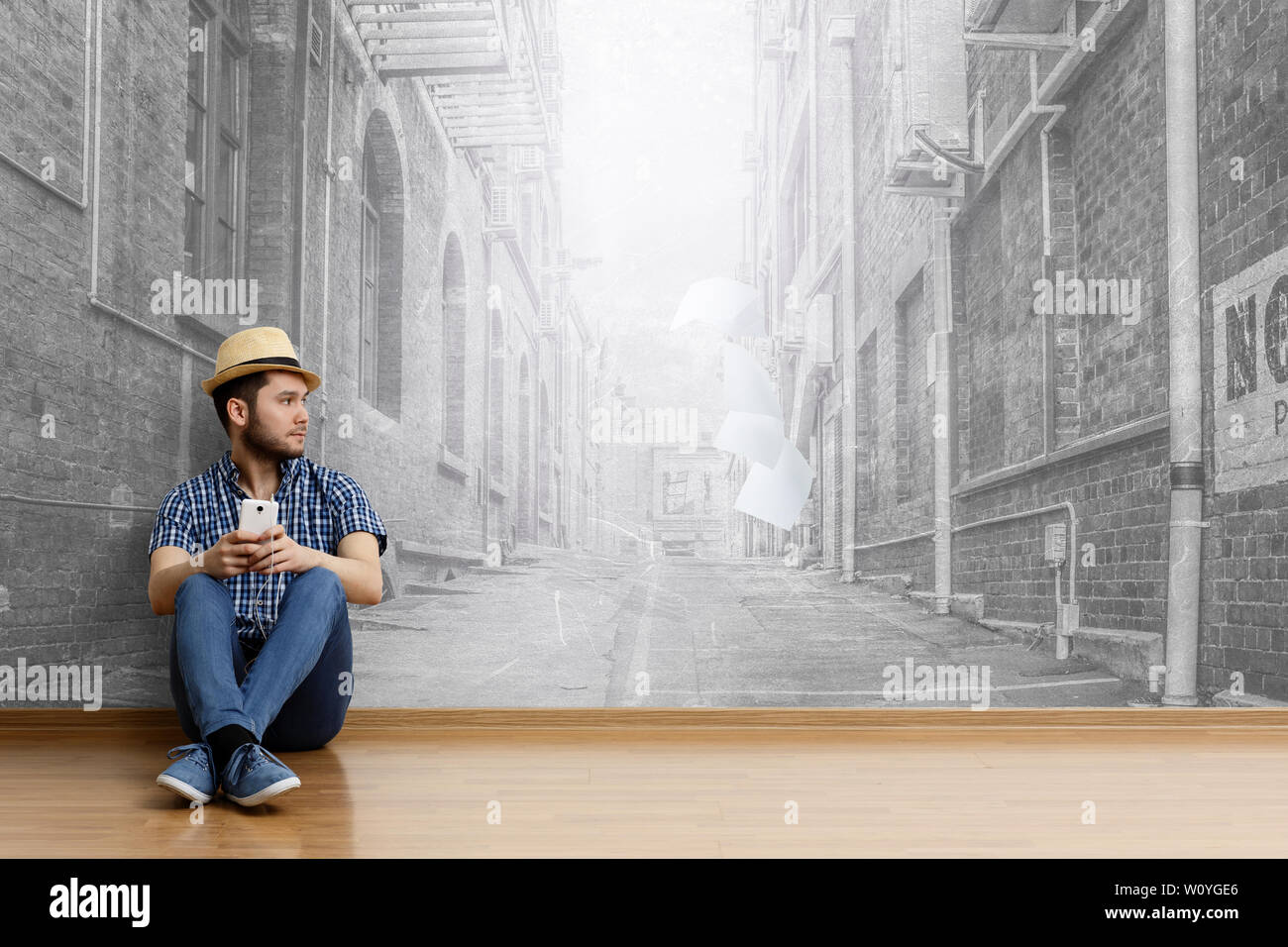 Junge fröhliche Mann in legere Kleidung und Hut sitzen auf dem Boden, stadtbild Hintergrund. Mixed Media Stockfoto