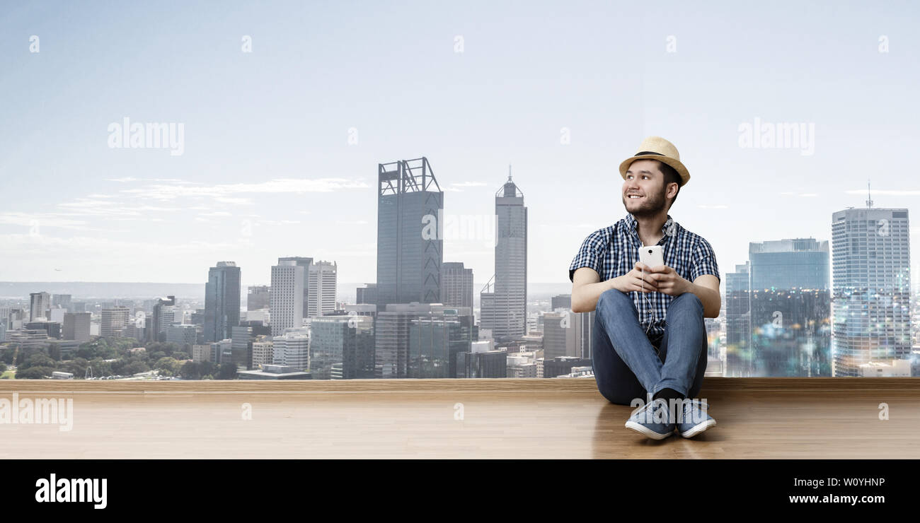 Junge fröhliche Mann in legere Kleidung und Hut sitzen auf dem Boden, stadtbild Hintergrund. Mixed Media Stockfoto