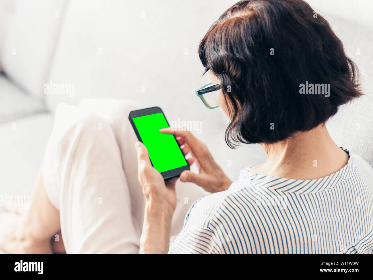 Im mittleren Alter brünette Frau mit Brille auf das graue Sofa Einsatz von Green Screen smartphone Stockfoto