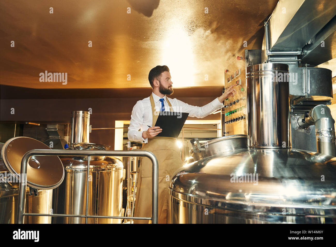 Stattliche Ingenieur der Brauerei Inspektion Prozess Bier brauen. Arbeitnehmer in Braun Schürze steht in der Nähe von Anlagen, Tanks, Ordner, Stockfoto