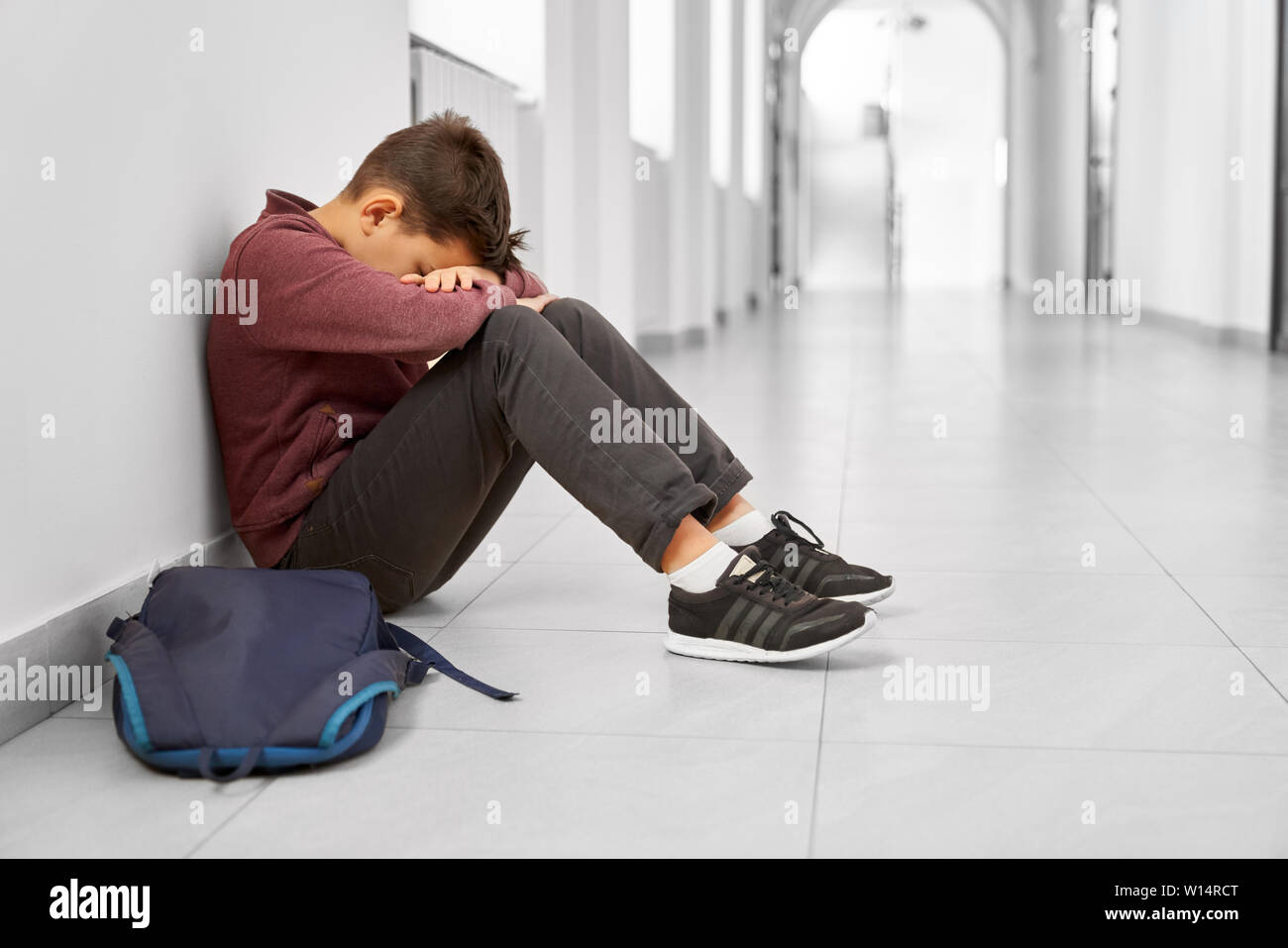 Seitenansicht des traurigen Schule Junge sitzt alleine auf dem Boden im Flur und verstecken Gesicht durch die Hände. Brunet Junge mit schlechten Mark und schrecklichen Stimmung nach dem Unterricht, zu weinen. Harte Zeit der Teenager und Studieren. Stockfoto