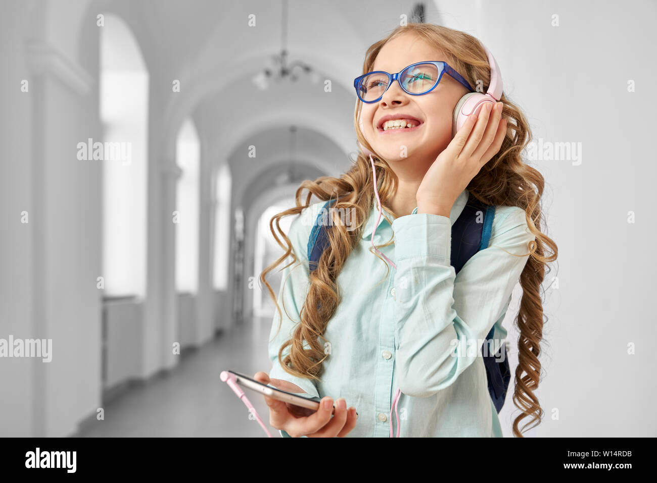 Lächelnd hübsches Mädchen tragen Brillen und Rucksack hören Musik von Big Pink Kopfhörer, weg schauen und Träumen. Funny Girl Ausruhen nach Lektionen, mit Smartphone und nach Hause gehen. Stockfoto