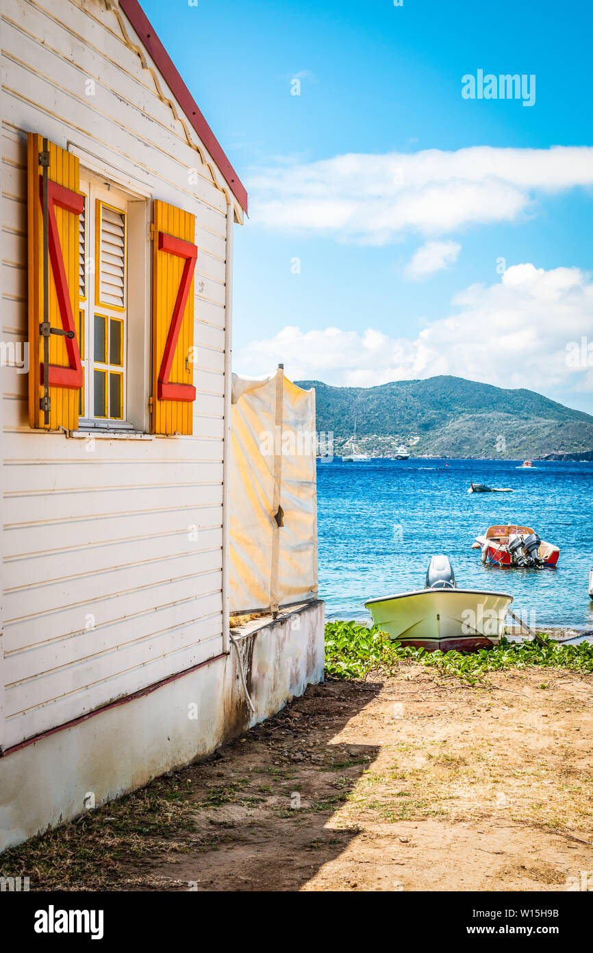Holz- Seitenwand eines weißen Haus mit gelben und roten Fensterläden. Im Hintergrund die Bucht von Wasser mit einem Motorboot am Strand. Terre-de-Haut, Stockfoto