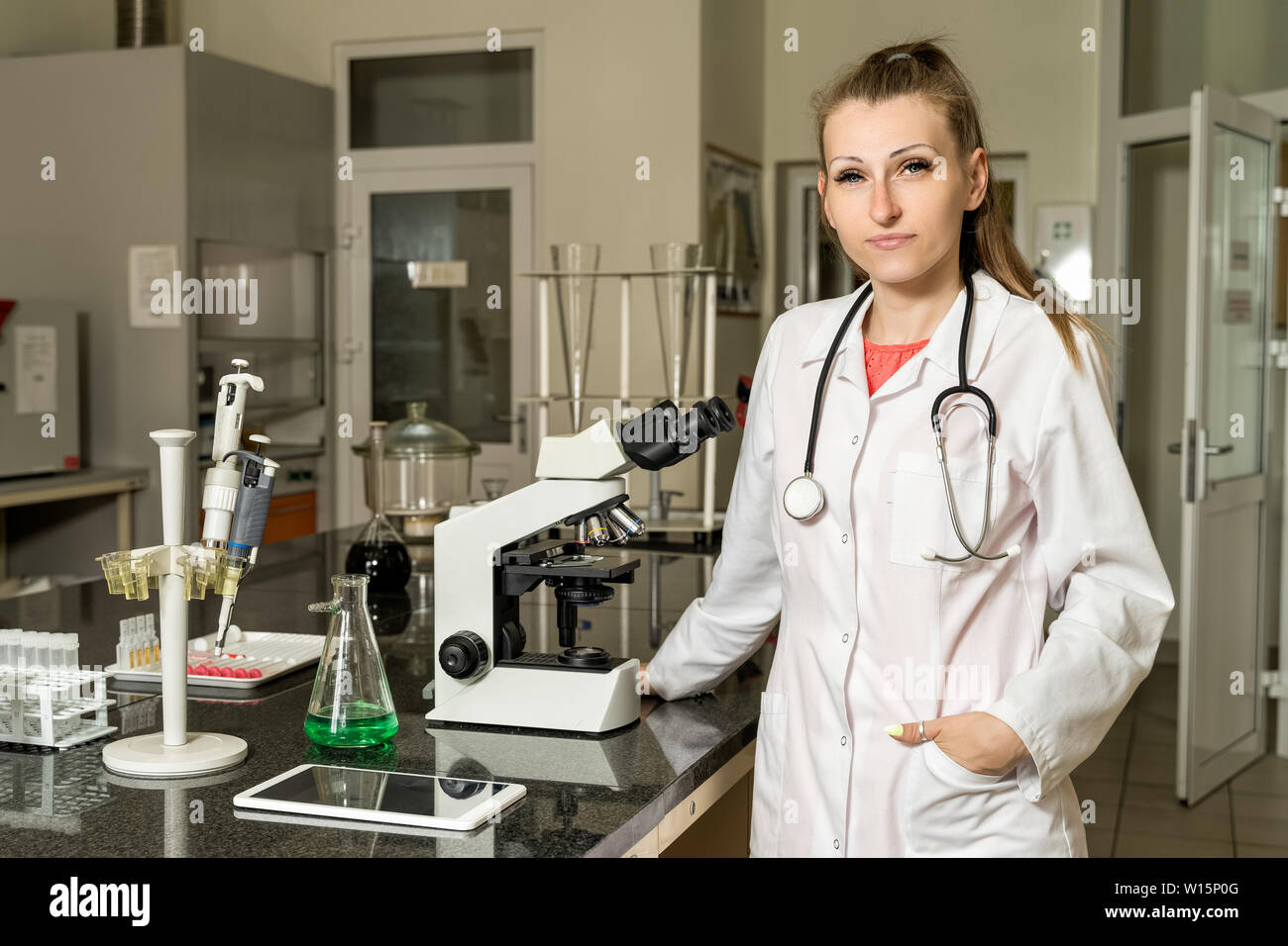 Junge, Kaukasier weiß, weiblich Labortechniker in eine weiße Robe mit Stethoskop neben Mikroskop-, Labor-, Geräte- und gl-Compound Stockfoto