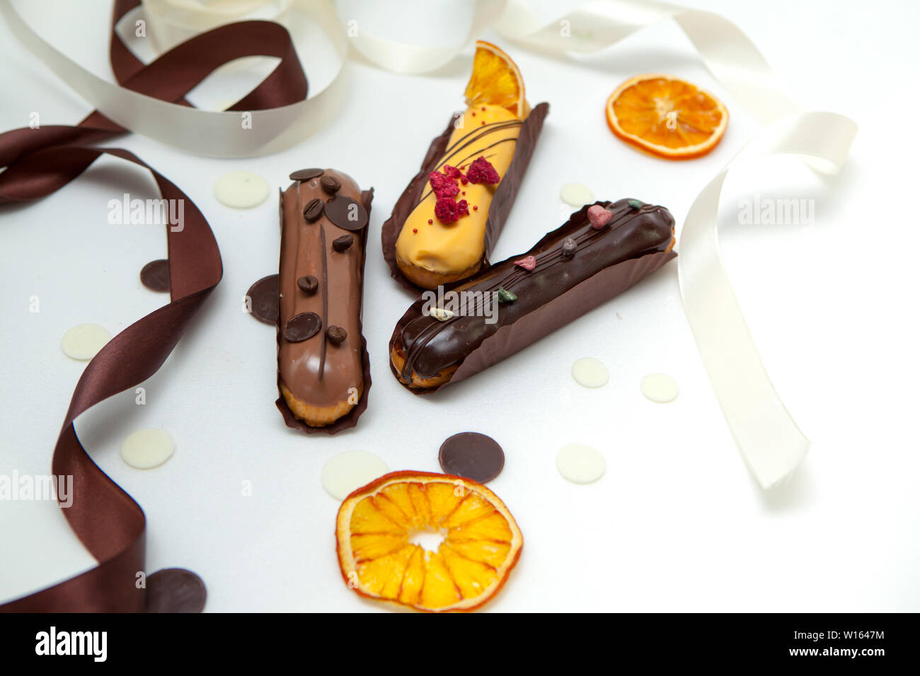 Eine Nahaufnahme von einem Satz von drei eclairs mit verschiedenen Füllungen und Design auf eine weiße Oberfläche mit Schokolade Tropfen eingerichtet, getrocknete Orangen und b Stockfoto