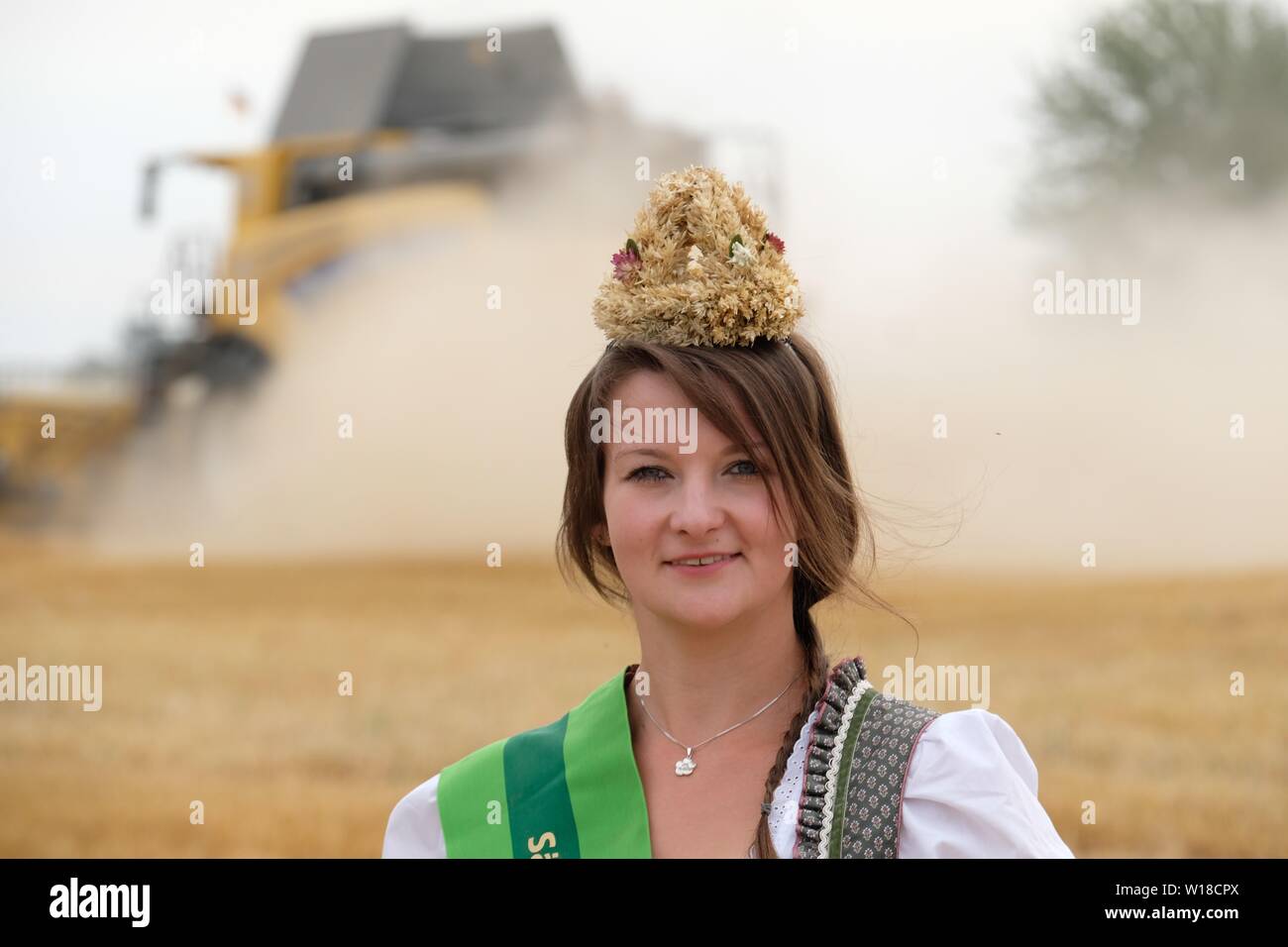 Grimma, Deutschland. 01. Juli, 2019. Elisabeth Dreher, Sachsens Ernte Königin 2019, steht auf einem wintergerste Feld während der Ernte Maschine im Hintergrund fahren ist. Die Landwirte in Sachsen befürchten eine erneute Dürre nach der Dürre von 2018. Credit: Sebastian Willnow/dpa-Zentralbild/dpa/Alamy leben Nachrichten Stockfoto