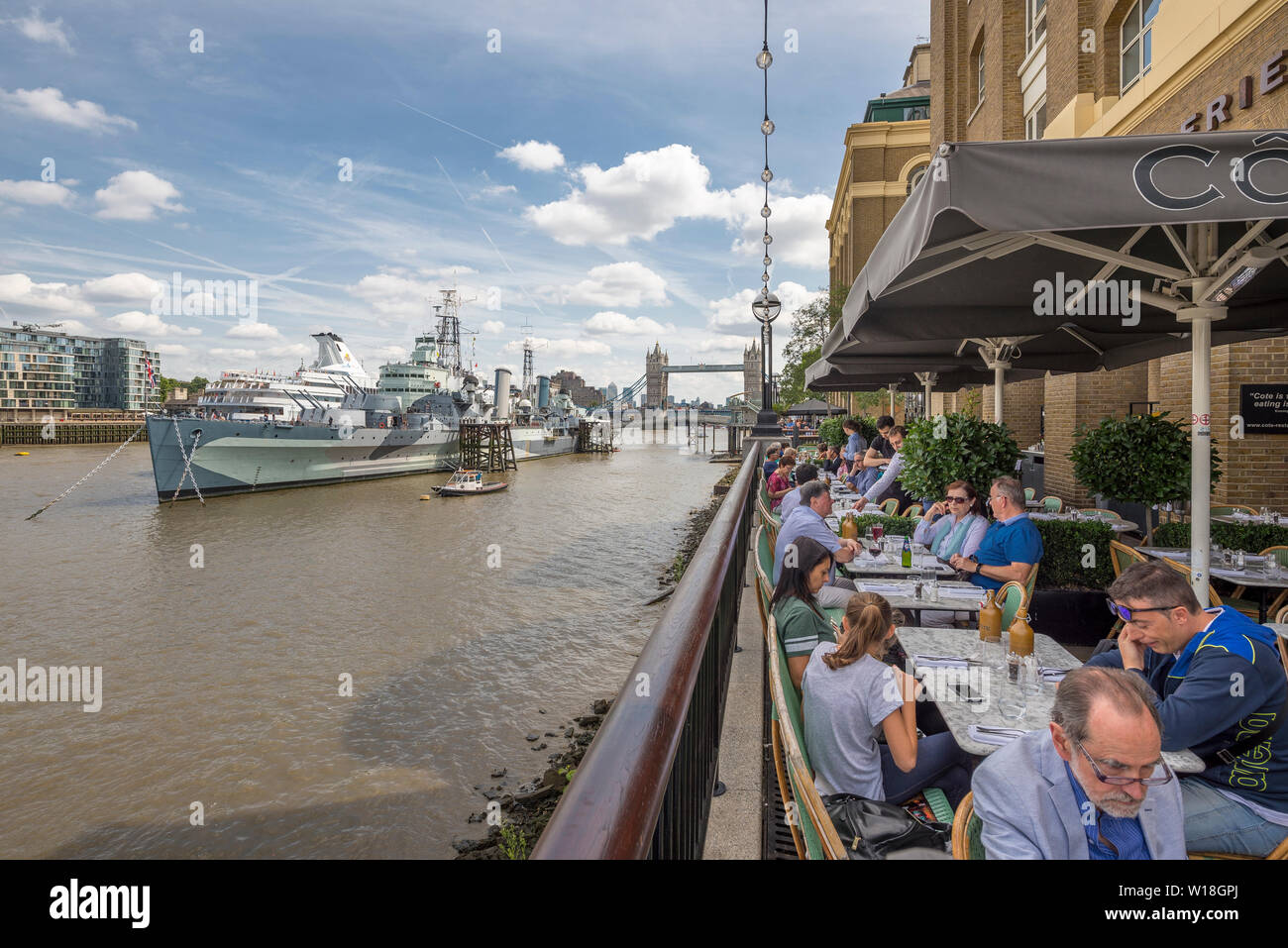 Eine belebte Restaurantterrasse mit Blick auf die themse und die HMS Belfast im Sommer im Touristengebiet an der South Bank in Central London Stockfoto