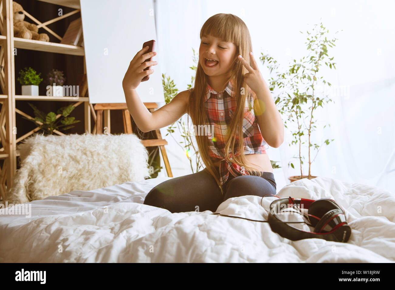 Mädchen über verschiedene Geräte zu Hause. Kleines Modell in ihrem Bett sitzend mit Smartphone und selfie oder über Videochat mit ihren Freunden. Konzept der Interaktion von Kindern und moderne Technologien. Stockfoto