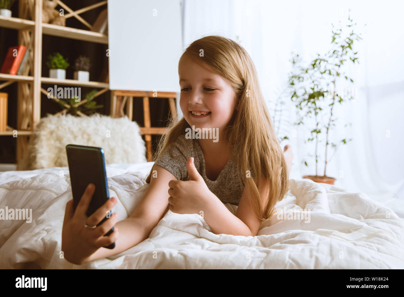 Mädchen über verschiedene Geräte zu Hause. Kleines Modell in Ihrem Zimmer mit Smartphone sitzen und selfie oder über Videochat mit ihren Freunden. Konzept der Interaktion von Kindern und moderne Technologien. Stockfoto