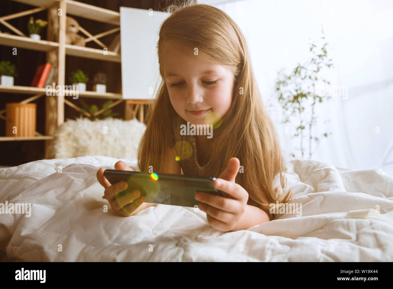 Mädchen über verschiedene Geräte zu Hause. Kleines Modell in Ihrem Zimmer mit Smartphone sitzen und selfie oder über Videochat mit ihren Freunden. Konzept der Interaktion von Kindern und moderne Technologien. Stockfoto