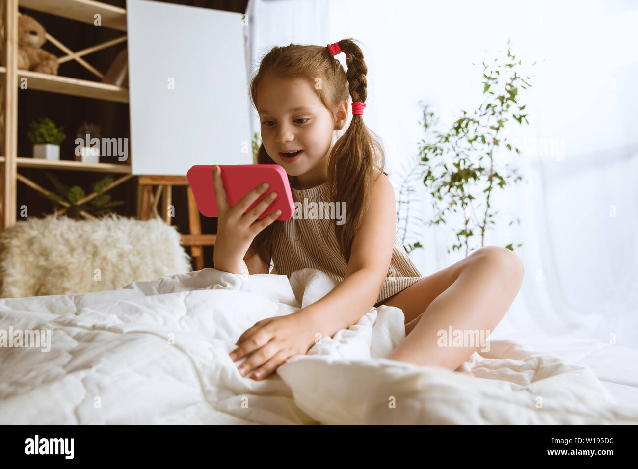 Mädchen über verschiedene Geräte zu Hause. Kleines Modell in Ihrem Zimmer mit Smartphone sitzen und selfie oder über Videochat mit ihren Freunden. Konzept der Interaktion von Kindern und moderne Technologien. Stockfoto