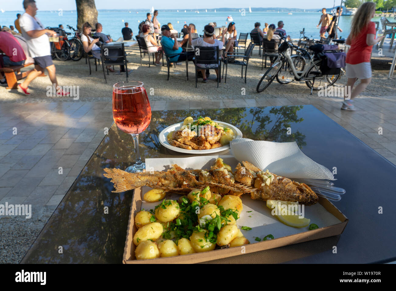 Balatonfüred, Ungarn, 06.10.2019: Fisch ad Weinfest gebratener Fisch Gerichte und ein Glas Rose Wein am Plattensee mit Menschen und den Hintergrund. Stockfoto