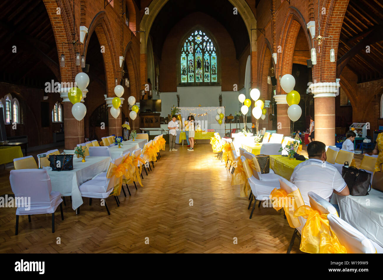 Eine Kirche Halle mit Tischen und Stühlen im weißen Stoff mit gelbem Band Bogen bereit für eine Party oder Rezeption gekleidet. Stockfoto