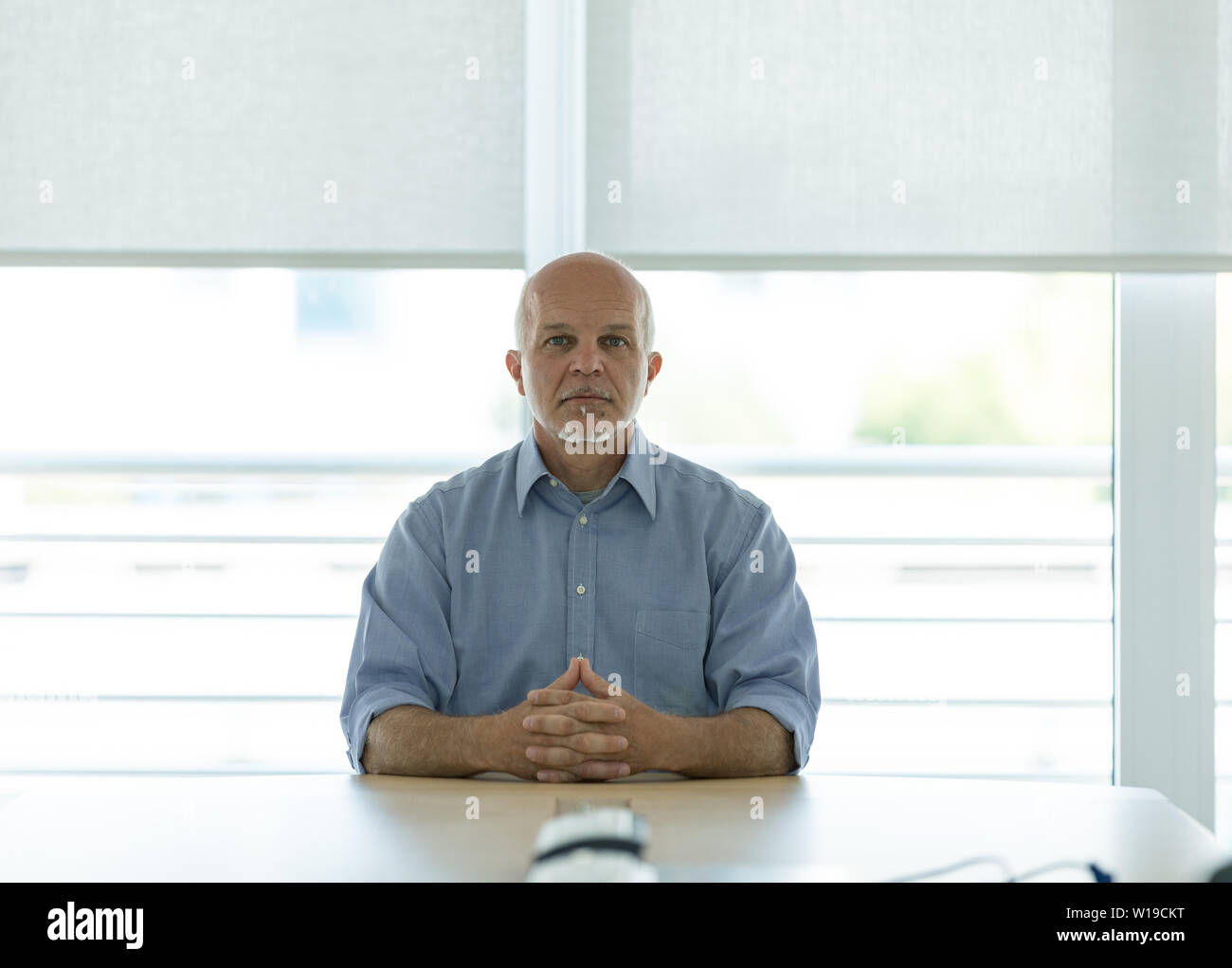 Senior Geschäftsmann saß am Schreibtisch im Büro mit ernsten Ausdruck, Fenster im Hintergrund. Stockfoto