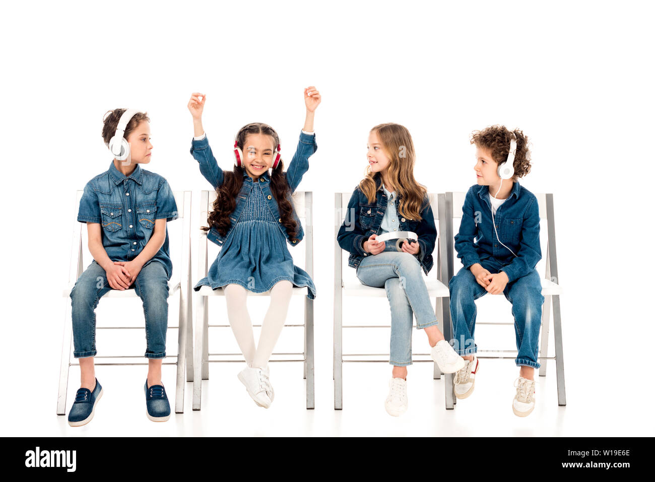 Vier Kinder in Jeans Kleidung sitzen auf Stühlen und das Hören von Musik über Kopfhörer auf Weiß Stockfoto